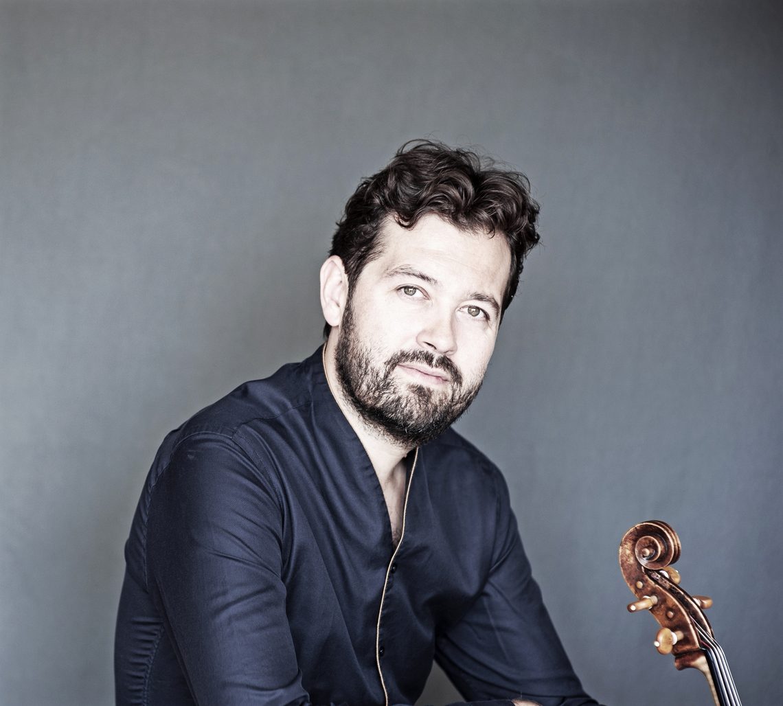 Main with curly black hair and beard wearing a blue jacket and holding a viola.