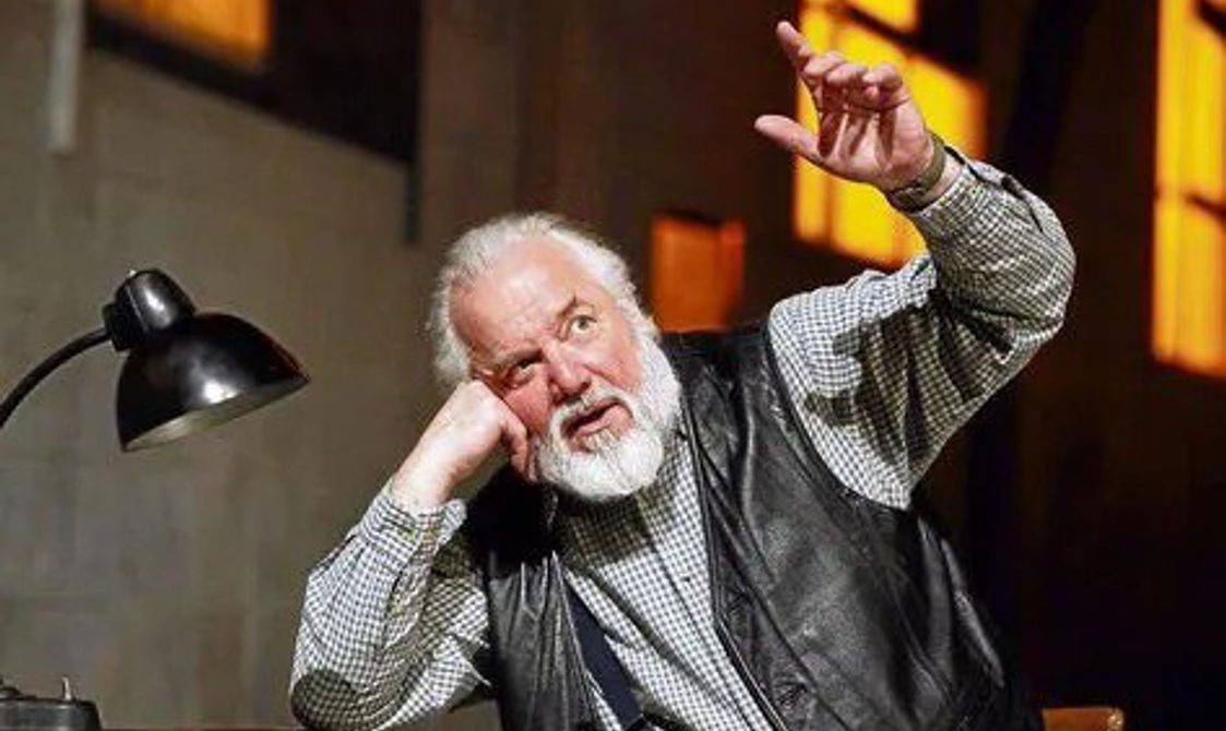 Man with white hair and beard sitting at a desk with his arm raised and singing.