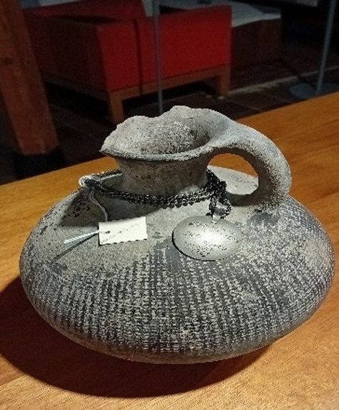 Ancient amphora in grey with ribbed curved body and small handle, and a small handwritten label attached.