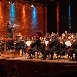 conductor onstage conducting an orchestra in a concert hall, illuminated by blue and red lights
