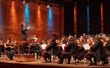 conductor onstage conducting an orchestra in a concert hall, illuminated by blue and red lights