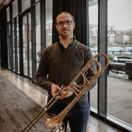 Man standing holding a brass instrument against a background of windows