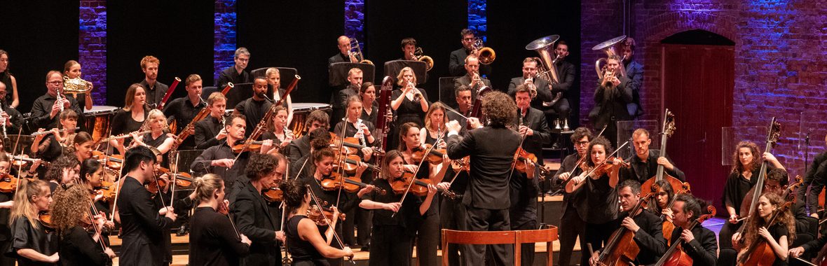 Orchestra onstage dressed in black playing instruments against a background of blue and purple lights