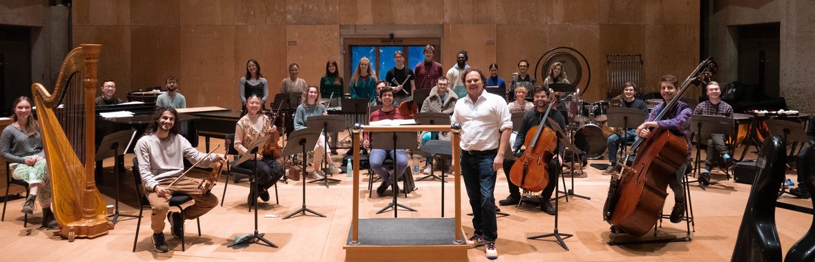 Musicians sitting with their instruments on a wooden floor, at the front the conductor stands beside his podium