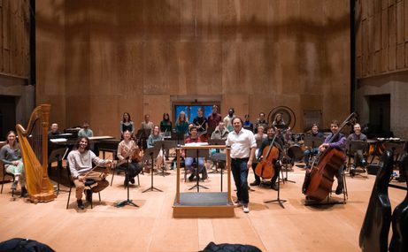 Musicians sitting with their instruments on a wooden floor, at the front the conductor stands beside his podium