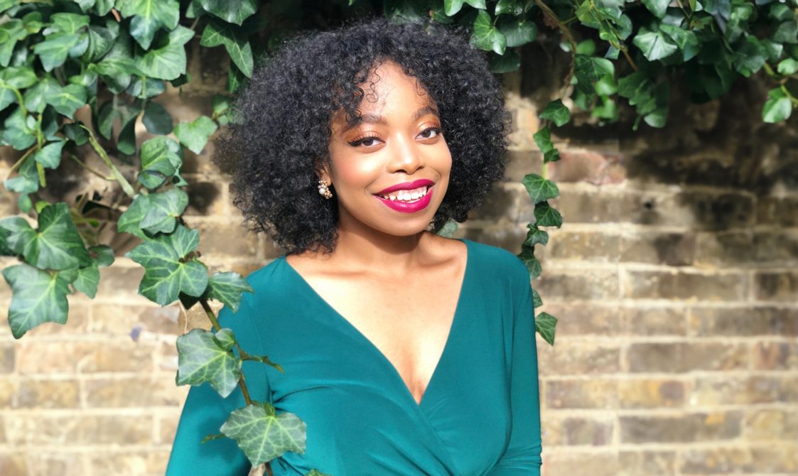 Woman with short black hair wearing a green dress and smiling, standing in front of a wall with green leaves hanging down.