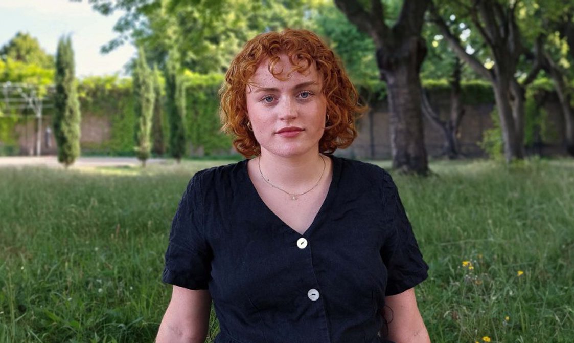 Woman with short red hair wearing a black top, standing outside with grass and trees behind her.