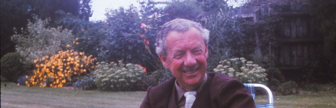 Man with grey hair smiling and sitting in a garden chair with trees and flowers behind him