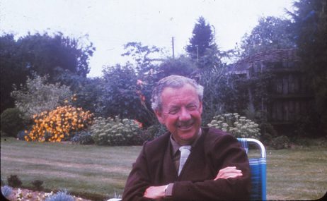 Man with grey hair smiling and sitting in a garden chair with trees and flowers behind him