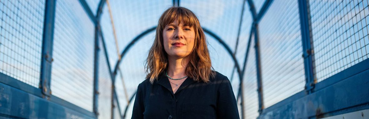 Woman with long hair wearing a black dress standing on a covered bridge, with blue sky behind her