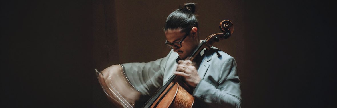 Musician seated playing the cello, his arm blurred by the movement, against a dark background