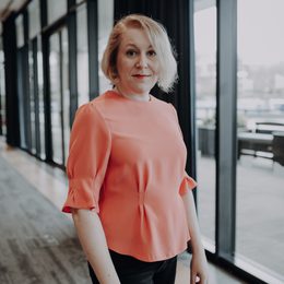Woman standing indoors in an orange top, against a background of windows