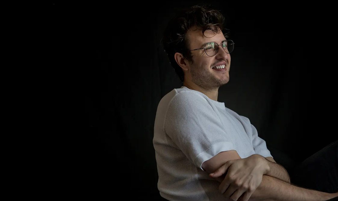 Man wearing a white t-shirt looking away from the camera and smiling, against a black background.