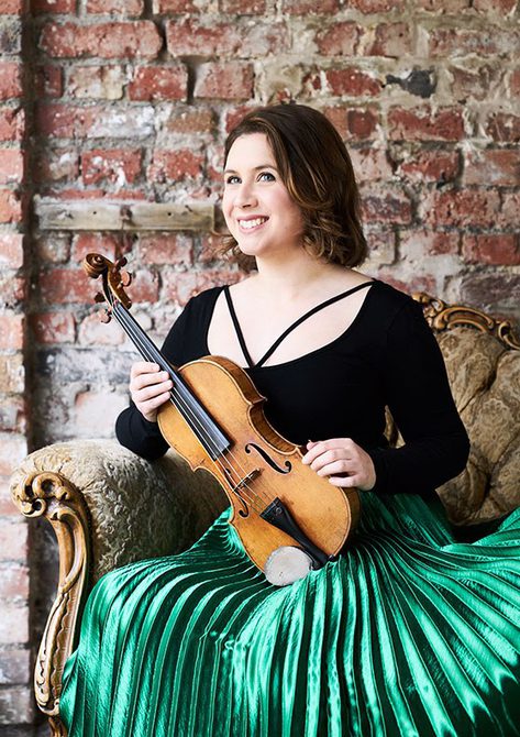 Woman with bobbed dark hair wearing a green pleated skirt, holding a violin and smiling.