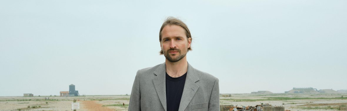 Man with dark hair and a beard, wearing a grey jacket, standing on a stretch of beach with tufts of grass and buildings in the distance.