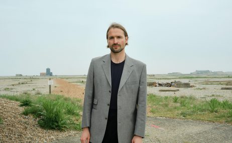 Man with dark hair and a beard, wearing a grey jacket, standing on a stretch of beach with tufts of grass and buildings in the distance.