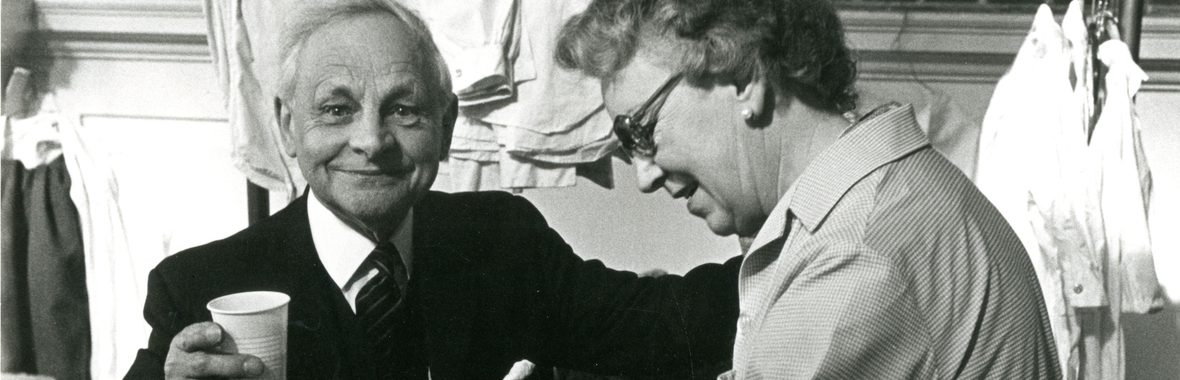 Black and white image of older man with white hair wearing a dark suit, standing beside a woman ironing a costume.