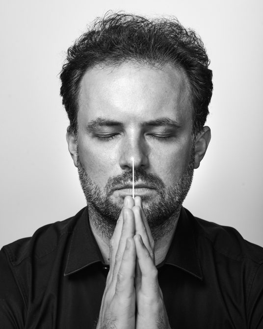 black and white close-up of a man with short dark hair and a beard, eyes closed, hands together.