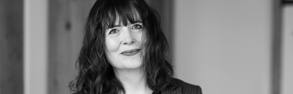black and white image of a woman with mid-length dark hair, wearing a jacket and smiling.