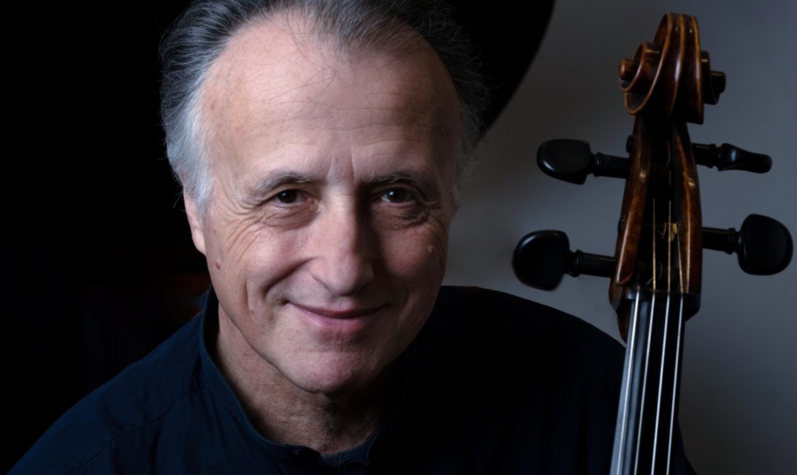 Man with grey hair smiling into the camera with the head of a cello beside him.