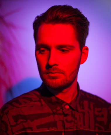 Close-up up of a man with short dark hair and a beard wearing a patterned top, lit with pink and purple light.