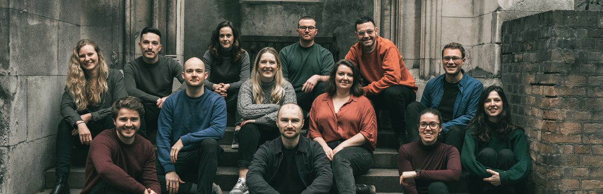 Sansara Choir, men and women wearing coloured clothing sitting on steps in a church and smiling.