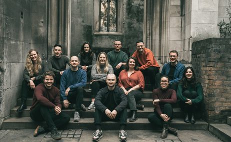 Sansara Choir, men and women wearing coloured clothing sitting on steps in a church and smiling.