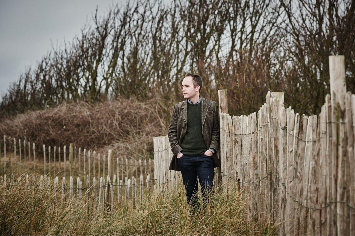 Man wearing a tweed jacket standing beside a fence amidst grass and woodland.