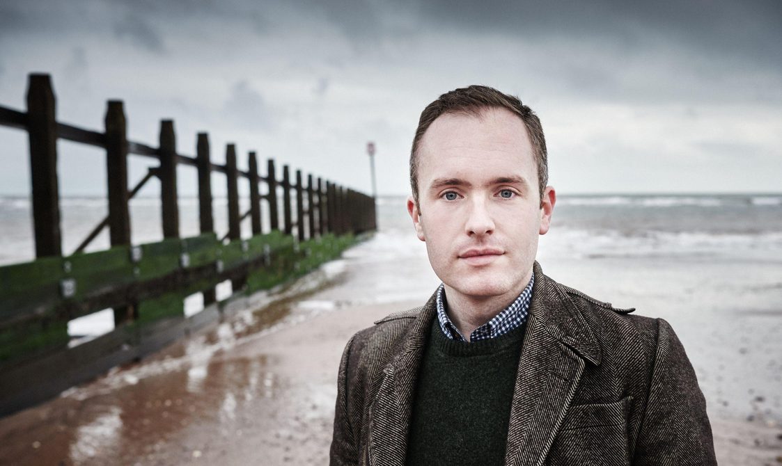 Man with short dark hair wearing a jacket, standing on a beach.
