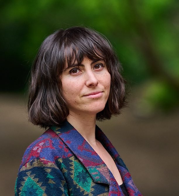 Woman with dark bobbed hair, wearing a patterned shirt and smiling, with blurred trees in the background.