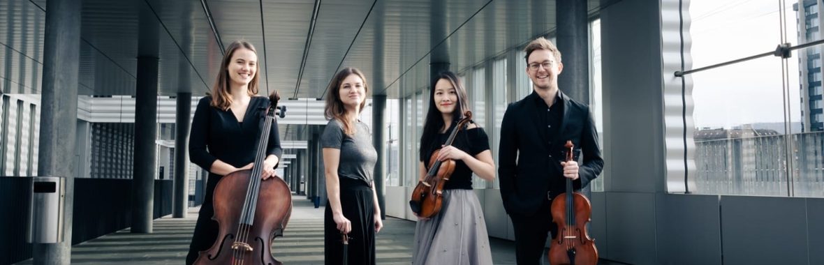Four musicians standing side by side holding instruments with a concrete building and windows behind them