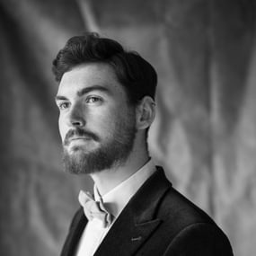 Man with dark hair and a beard wearing a black jacket and bow tie, looking into the distance.