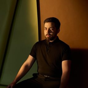 Man with dark hair and beard wearing a black tshirt sitting in a dark room.
