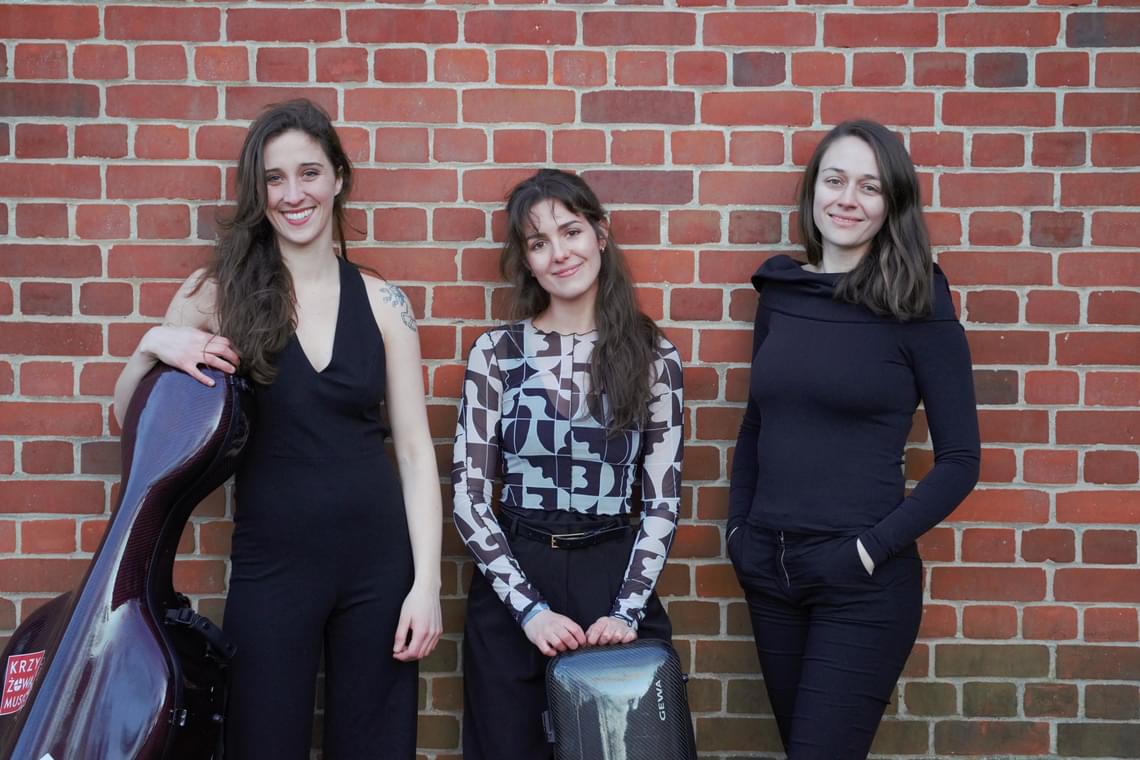 Three women standing together against a brick wall, two of them holding instrument cases.