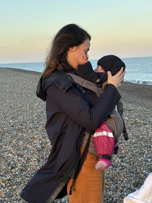 Woman wearing a rain jacket and carrying a baby on the beach.