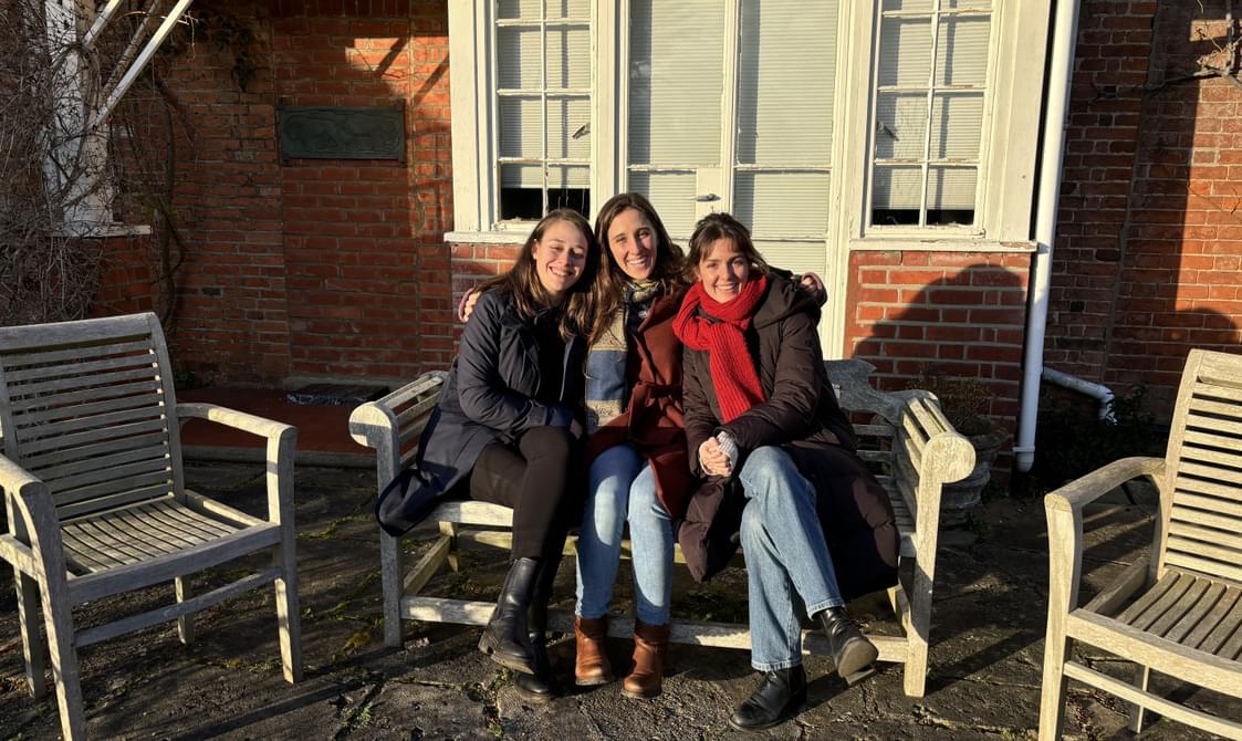 Three women sitting together on a bench outside and smiling.