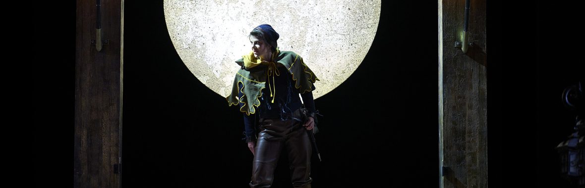 Performer onstage wearing medieval costume and cap with a full moon behind them.