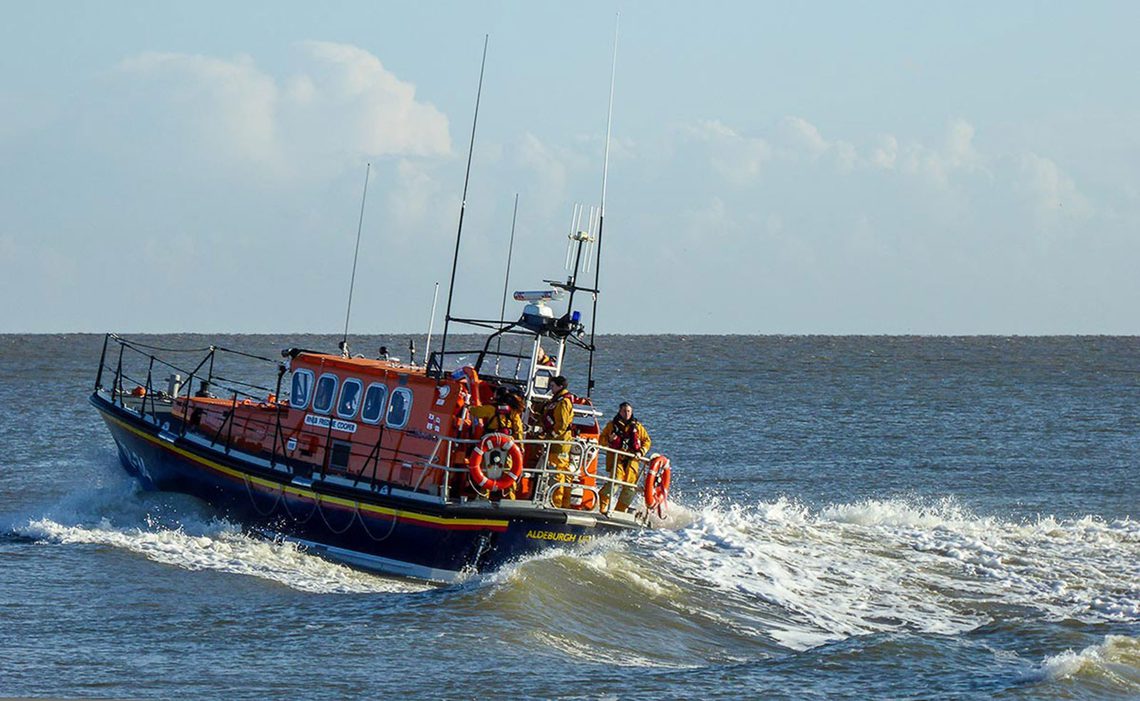 Orange and blue lifeboat cresting a wave on the sea with lifeboat men standing dressed in yellow at the stern of the boat