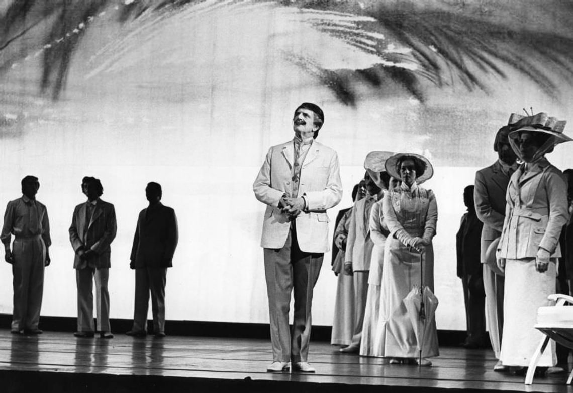 Black and white photo pf characters on stage, men in suits, woman in a wide-brimmed hat