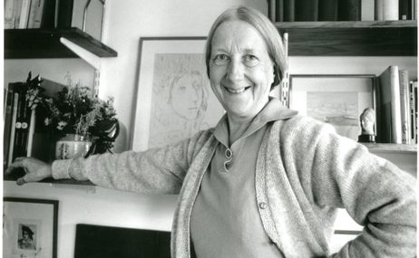 black and white image of a woman smiling and standing in a room filled with paintings and books.