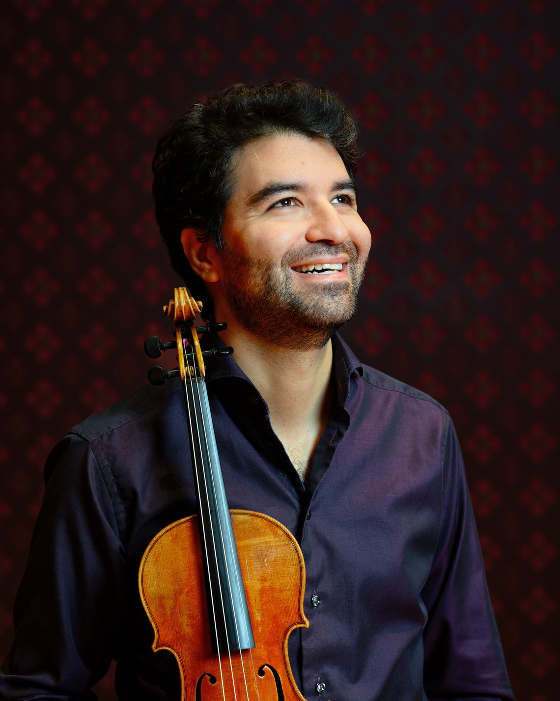 Man with dark hair and beard looking up and smiling, holding a string instrument