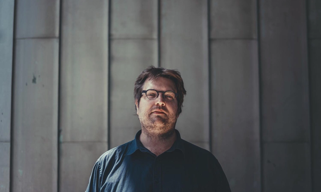 Man with dark hair and glasses standing against a concrete wall and looking at the camera.
