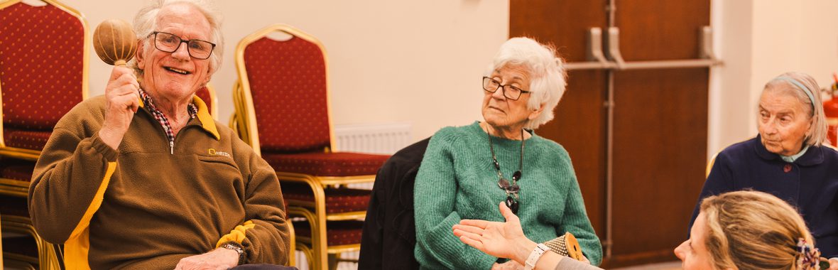 Man sitting in a chair holding a maraca and smiling, another woman is sitting beside him and holding out her hand to him