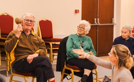 Man sitting in a chair holding a maraca and smiling, another woman is sitting beside him and holding out her hand to him