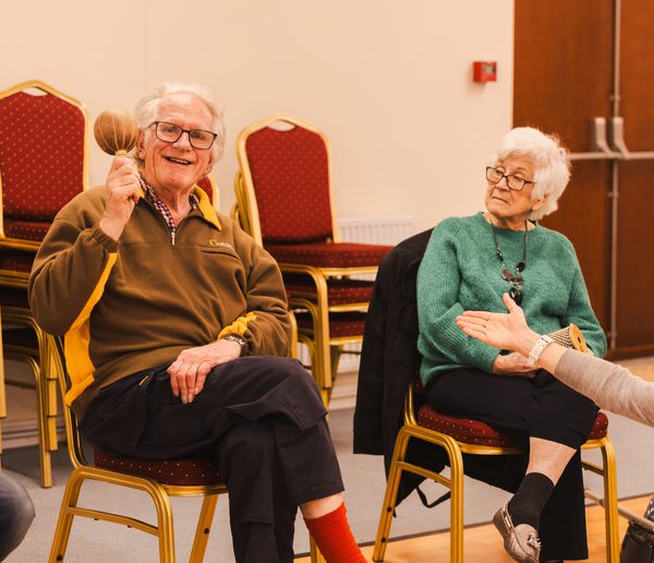Man sitting in a chair holding a maraca and smiling, another woman is sitting beside him and holding out her hand to him