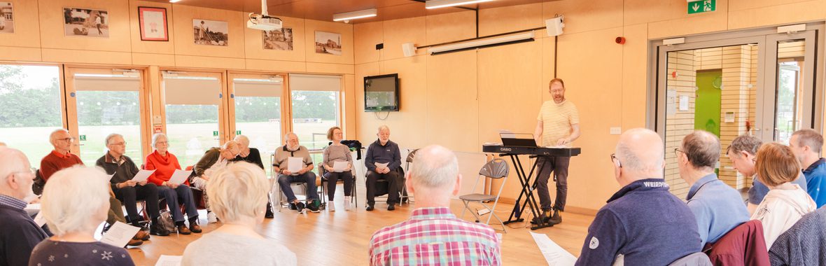 Group of older people sitting in a circle facing a man playing the keyboard and singing