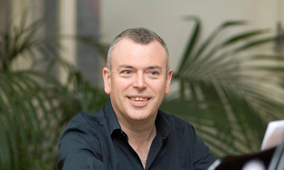 Grey haired man wearing a blue shirt looking at the camera, with fern plants behind him.