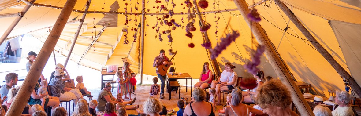 man playing the guitar in a large tent, with children and adults seated while watching him perform