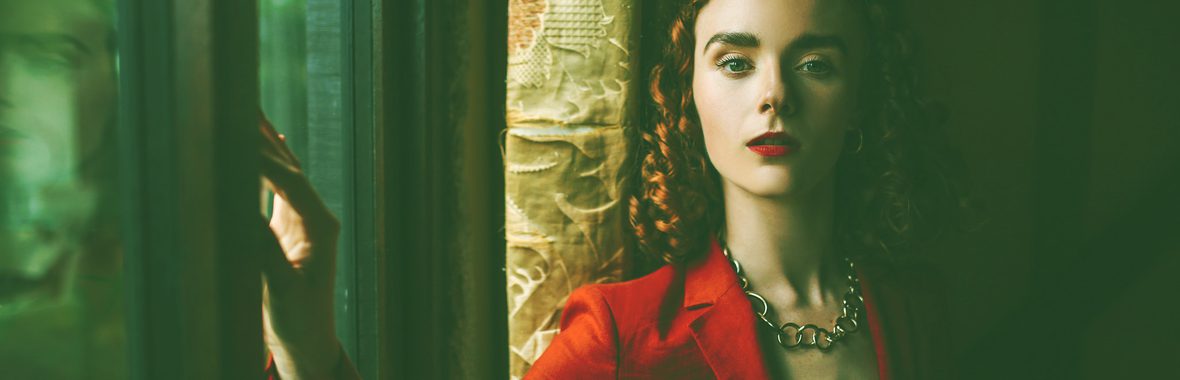 A woman in a red blazer leaning against a window, looking into the camera.