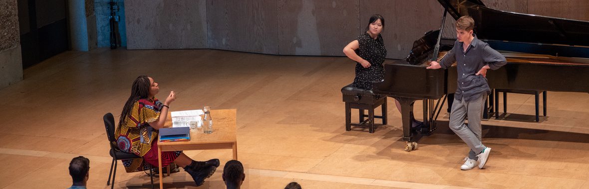 A woman sitting at a desk gesturing to two musicians leaning and sitting at a piano.